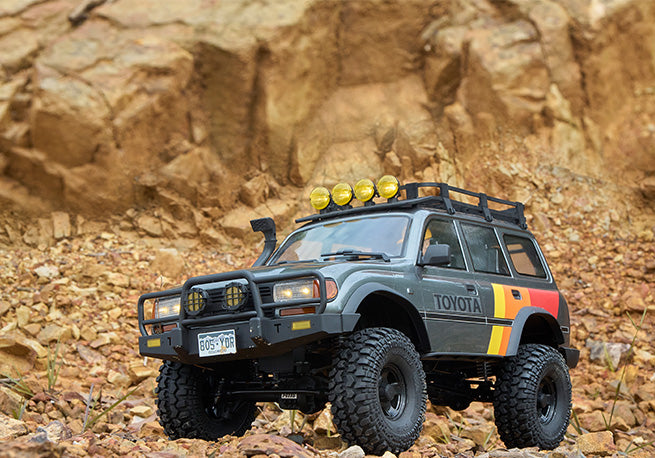 A detailed miniature Toyota SUV model with off-road modifications, including roof-mounted lights, a roof rack, and rugged tires, positioned on a rocky terrain with a natural cliff in the background.