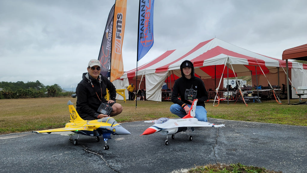 Two people kneeling next to yellow and white remote control model jets on an outdoor runway with a red and white striped tent and flags in the background.