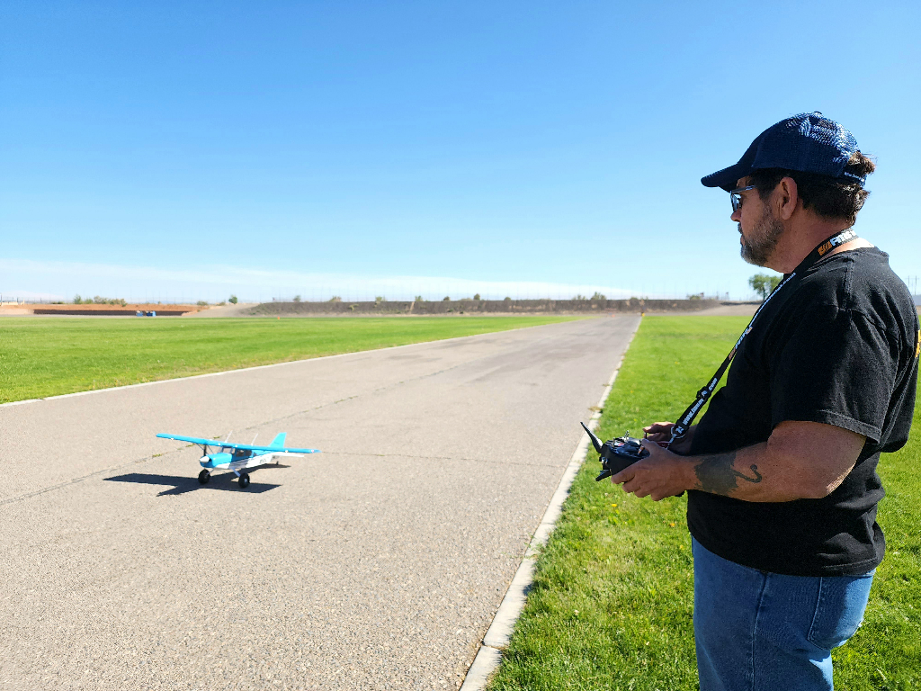 Person standing on the edge of a paved runway holding a remote control, operating a blue and white remote control model airplane on the runway with green grass and clear blue sky in the background.