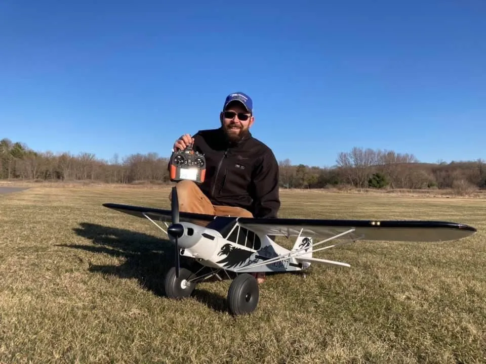 A man kneeling on a grassy field holds a remote control next to a large RC airplane.