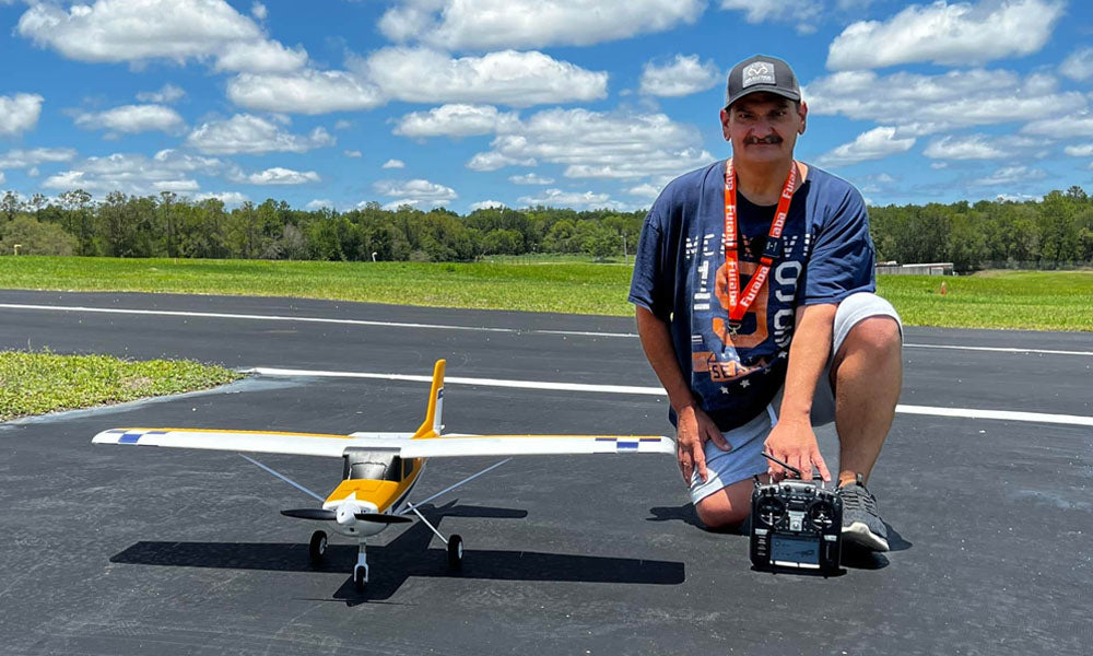 A hobbyist kneeling on a runway with his yellow and white RC model airplane, preparing for flight with a remote transmitter