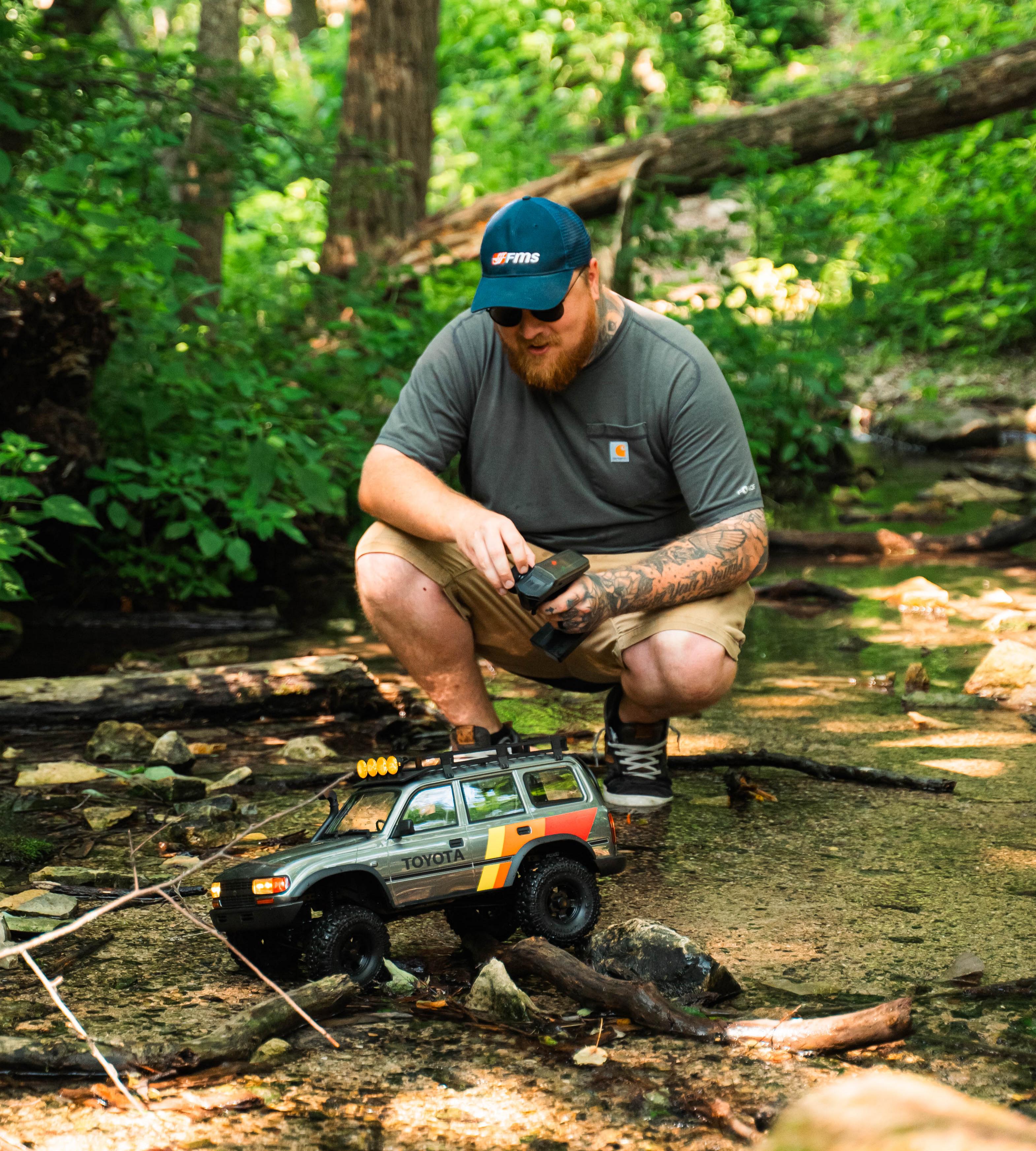 Man operating an FMS Toyota RC crawler model in a shallow outdoor stream.