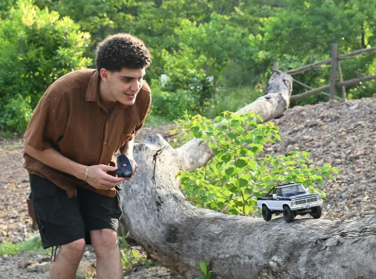 Hobbyist driving a hobby-grade RC crawler along a fallen log on a wooded trail using a pistol-grip transmitter
