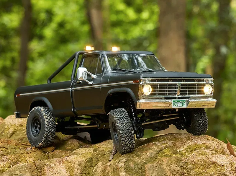 Matte black Ford pickup RC crawler with roll bar and working LED lights posed on a mossy forest rock