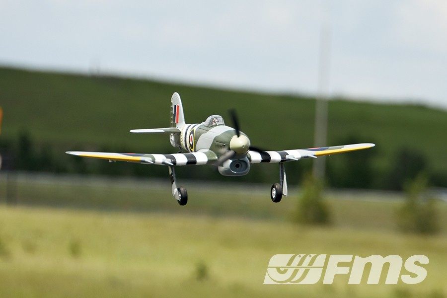 FMS 1100mm Typhoon PNP model airplane in flight, featuring green and grey camouflage with black and white stripes on wings.
