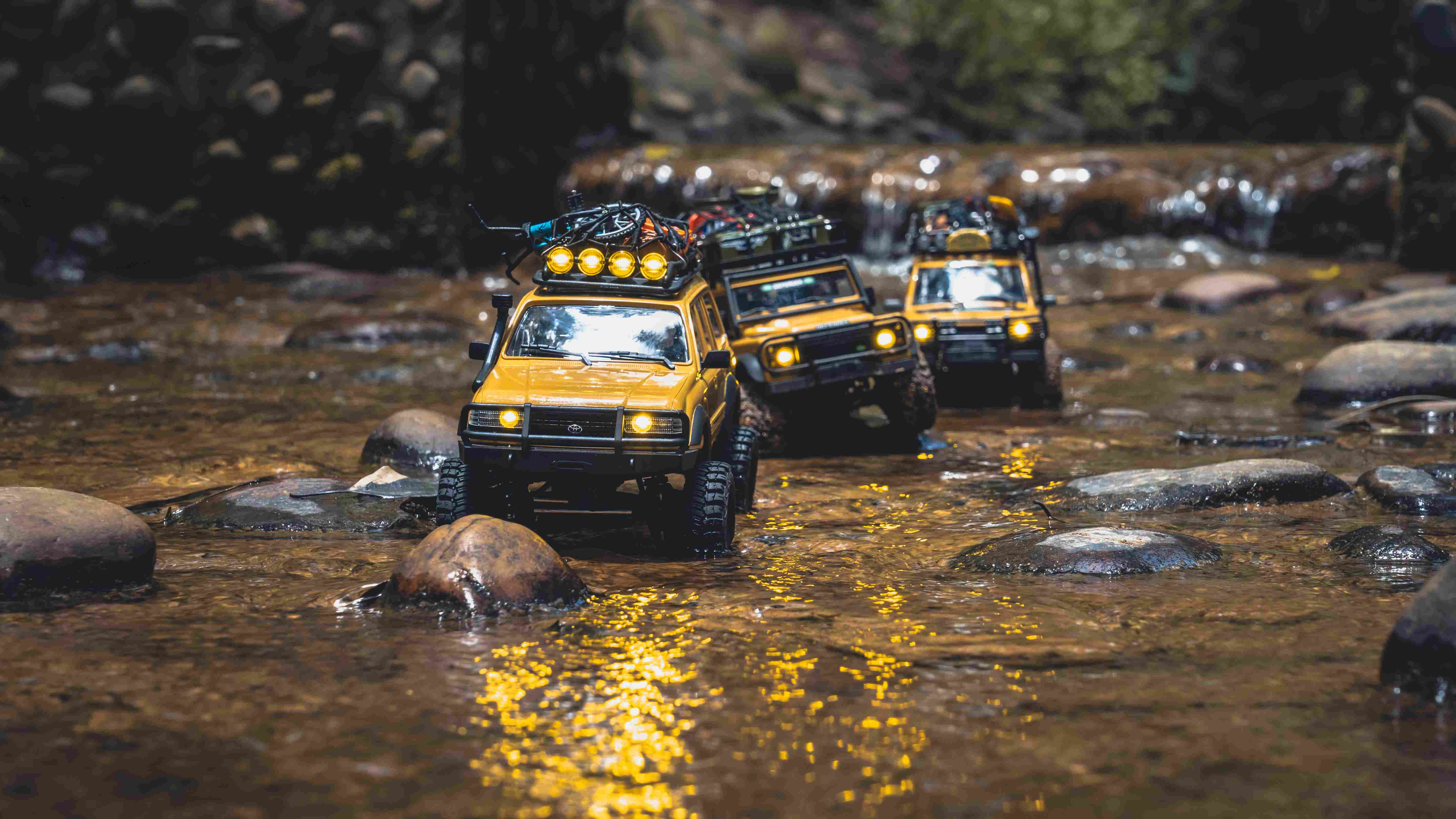 Three yellow FMS mini RC crawlers driving through a shallow rocky stream