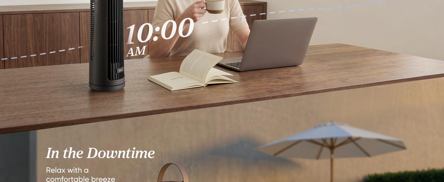 A person sits at a wooden table with a laptop, a book, and a cup, next to a black fan, in a relaxed indoor setting.