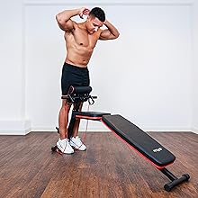 Adjustable workout bench with red trim. Person demonstrating its use for abdominal exercises on wooden floor against white wall.
