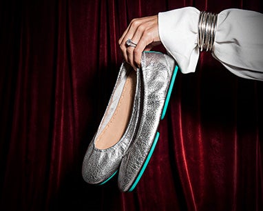 A hand adorned with silver bangles holds a pair of metallic silver Tieks flats against a deep red velvet backdrop.