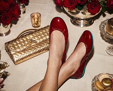 A woman lounges at an elegant holiday table wearing red patent Tieks flats, surrounded by red roses, gold accents, a quilted gold clutch, candles, and macarons.