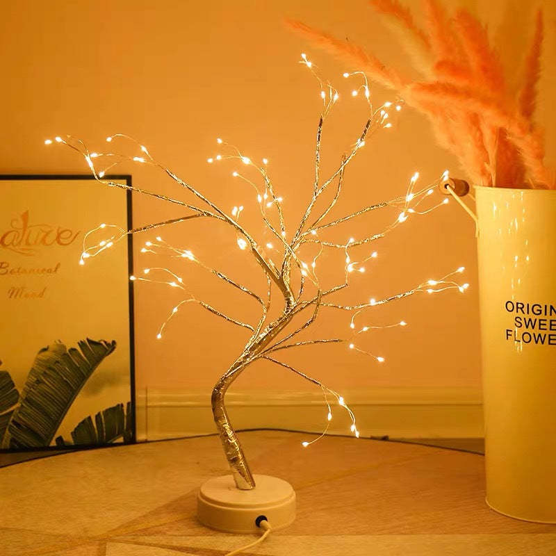 Decorative LED tree on a table with a warm glow, next to a vase with flowers and a framed picture.