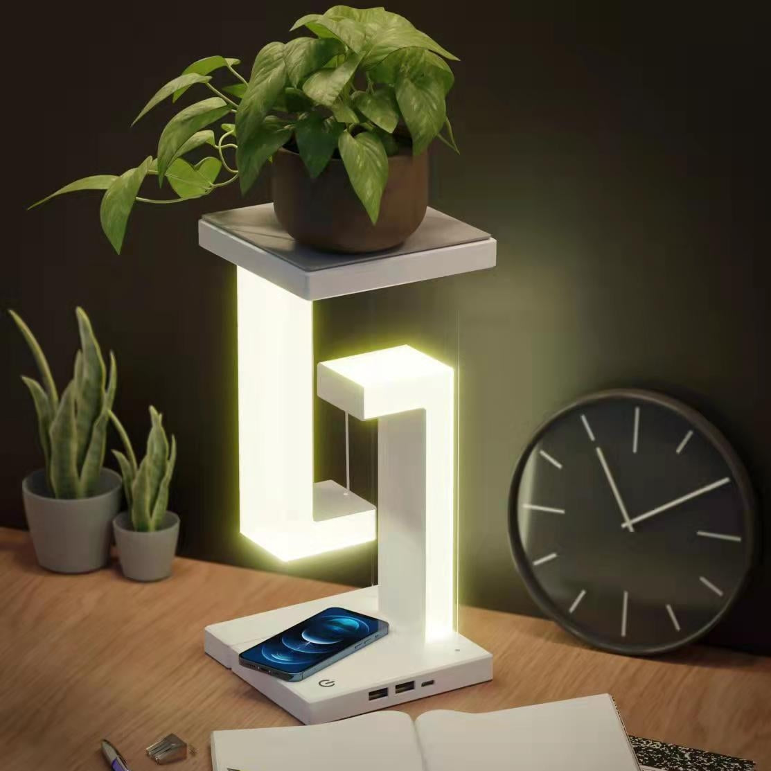 Modern desk lamp with phone charging station on a wooden surface with plants and a clock in the background.