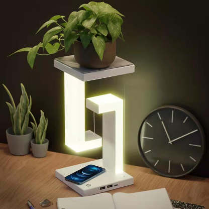 Modern desk lamp with phone charging station on a wooden surface with plants and a clock in the background.