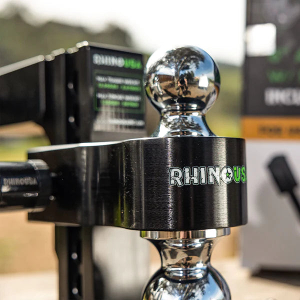 Close-up of a shiny metal trailer hitch ball mount with the brand name RHINO USA engraved on it. The background is blurred, showing outdoor scenery and equipment boxes.