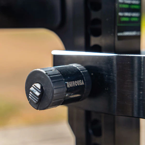 Close-up of a black Rhino USA hitch pin lock securing a metal trailer hitch. The lock is cylindrical with textured grip and branding, attached to shiny silver metal with a blurred outdoor background.