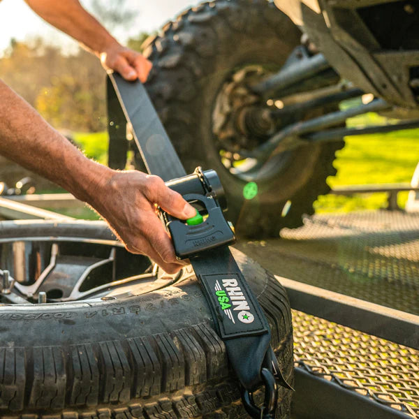 A person secures a large off-road vehicle tire with a Rhino USA ratchet strap on a metal trailer, fastening the tire for safe transport outdoors in daylight.