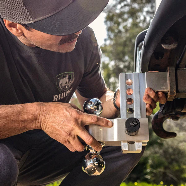 A man wearing a cap and a Rhino T-shirt installs a silver adjustable trailer hitch with dual chrome balls onto the back of a vehicle outdoors.