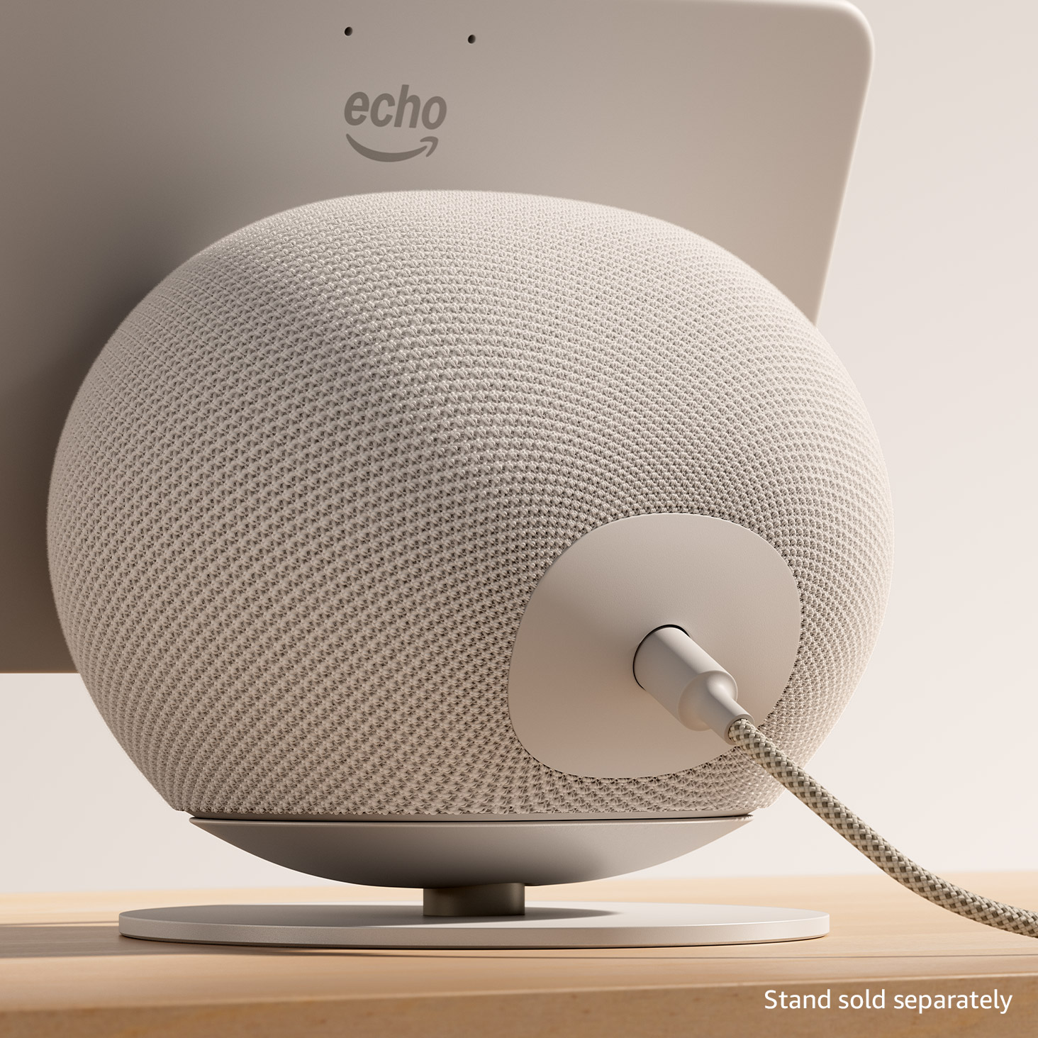 White Amazon Echo device, a spherical smart speaker with textured mesh covering, sitting on a small stand on a wooden surface. Echo logo visible on back panel with power cable connected. Text reads: "Stand sold separately.