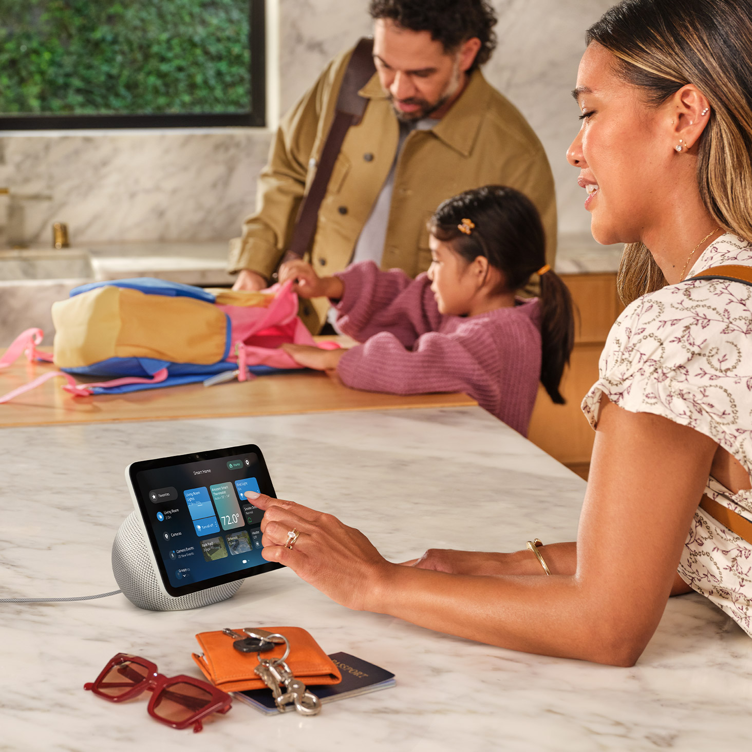 A family in a modern kitchen. A person uses an Echo Show 8 on a marble countertop, displaying a smart home interface. In the background, two people help a child with a colorful backpack.