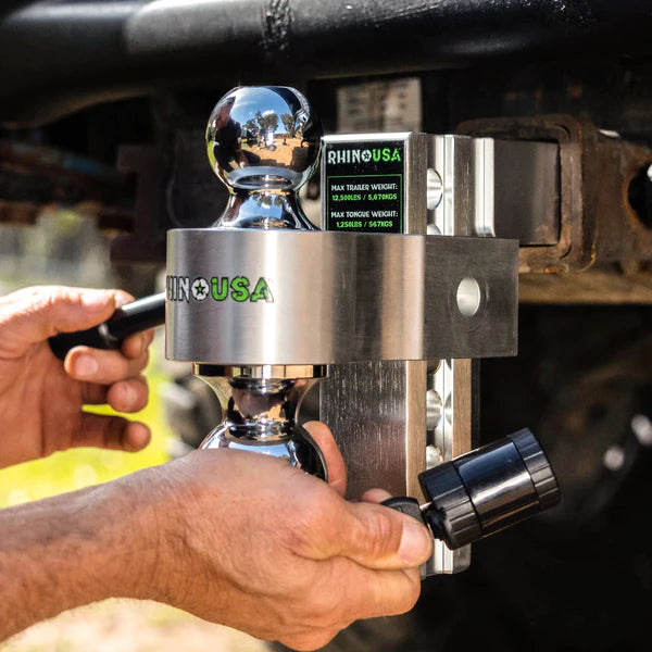 A person secures a shiny metal trailer hitch lock from Rhino USA onto a vehicle&rsquo;s hitch receiver, tightening it with a key outdoors in daylight.
