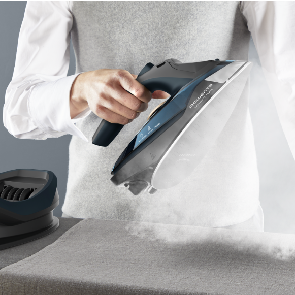 Hand using a Rowenta steam iron on a garment, releasing visible steam while pressing fabric on an ironing board.