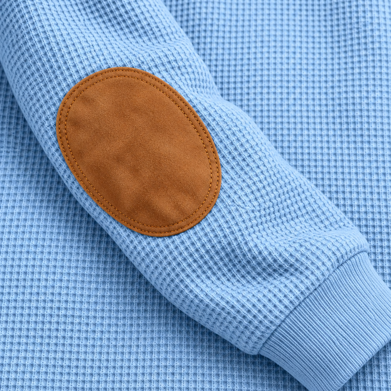 Close-up of a warm brown oval elbow patch with perimeter stitching on a Light Blue waffle-knit sleeve