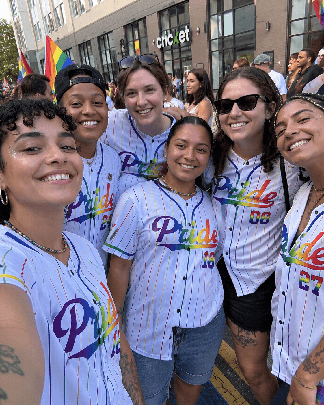 A close-up group photo of smiling friends celebrating, wearing custom white LGBTQ pride baseball jerseys.