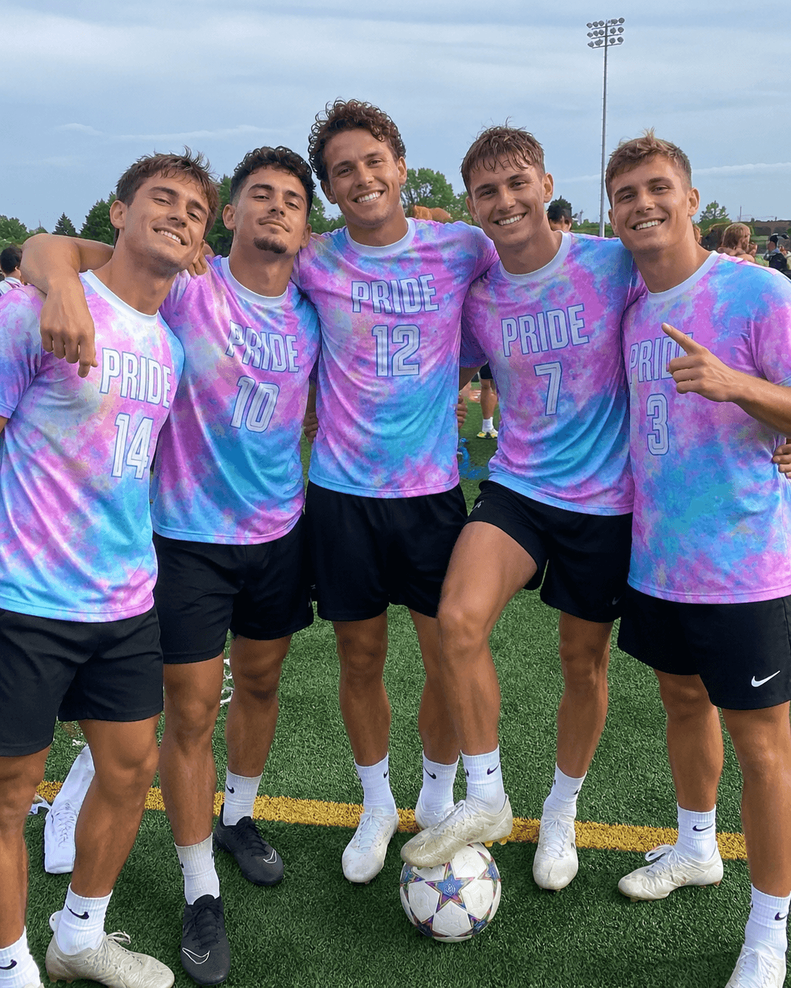 A group of male friends posing on a soccer field, wearing matching pink and blue tie-dye pride soccer jerseys.