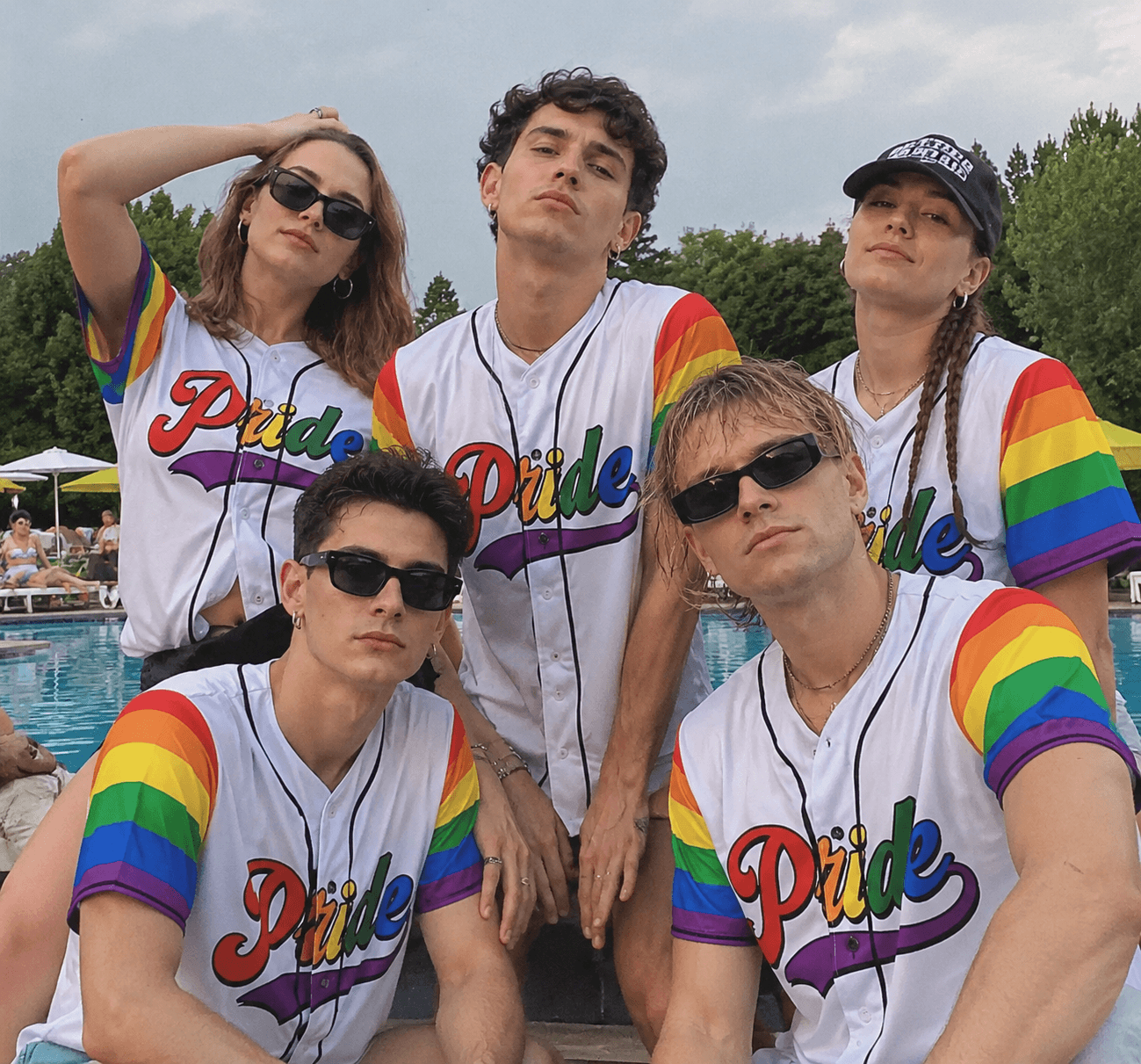 A group of four diverse friends laughing together outdoors, wearing matching white baseball jerseys with rainbow trim.