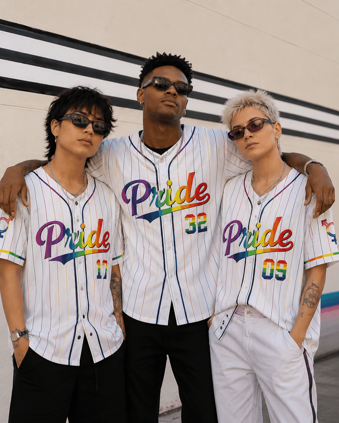 Three friends posing together in front of a wall, showcasing unisex white pride baseball shirts with rainbow accents.