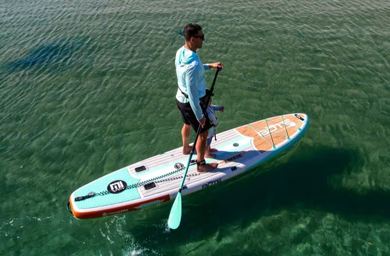 Man paddleboarding on clear water with a child seated at the front of the board.