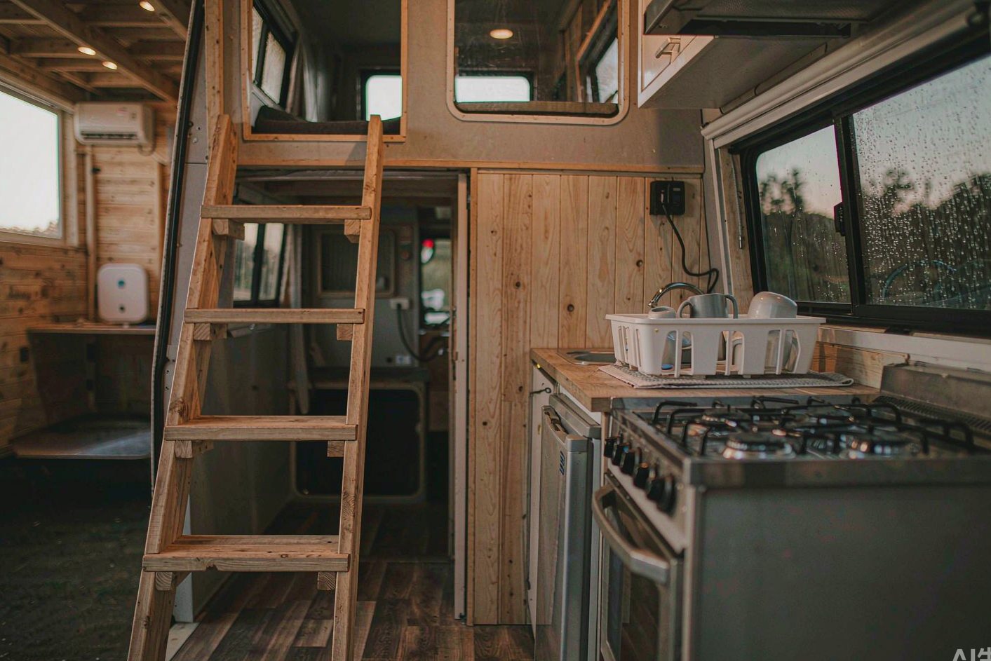 Compact American tiny house interior at dusk with a loft sleeping area, ladder, and small kitchenette with a propane cooktop, a small plain white rounded device resting far away in soft focus on a shelf, no logos, no people
