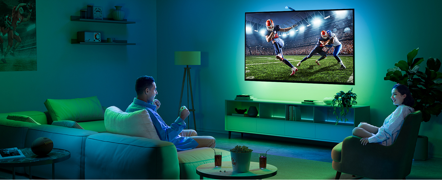 Large flat-screen TV mounted on wall, displaying football game. Two people watch from a couch in a dimly lit living room with ambient green lighting.