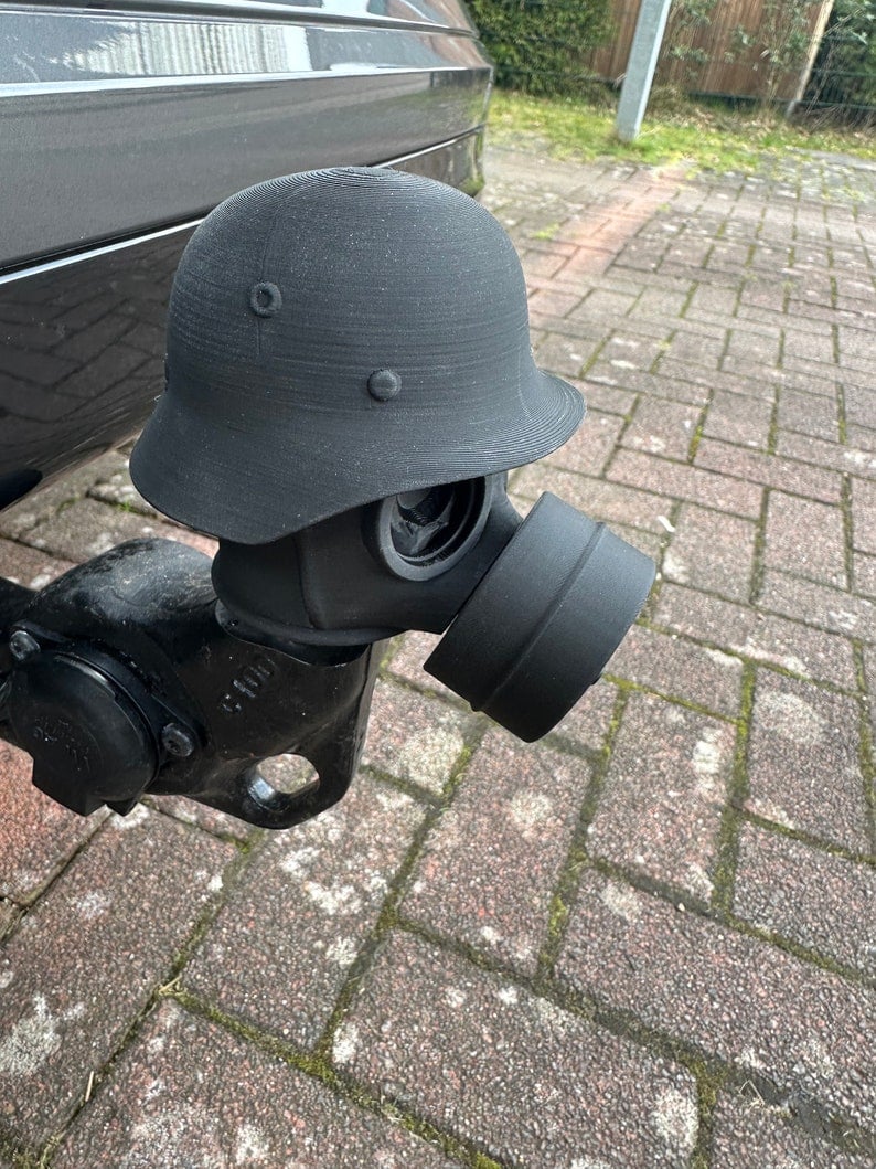 May include: A black gas mask attached to a black helmet. The helmet has a round shape with a brim and is attached to the gas mask with a black fabric covering the connection. The gas mask has a round filter attached to the front.