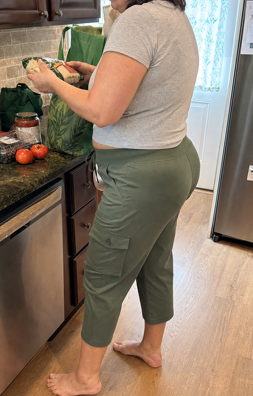 Camille, a plus-size woman, wearing Moss Olive stretch cargo capri pants while unpacking groceries in a family kitchen