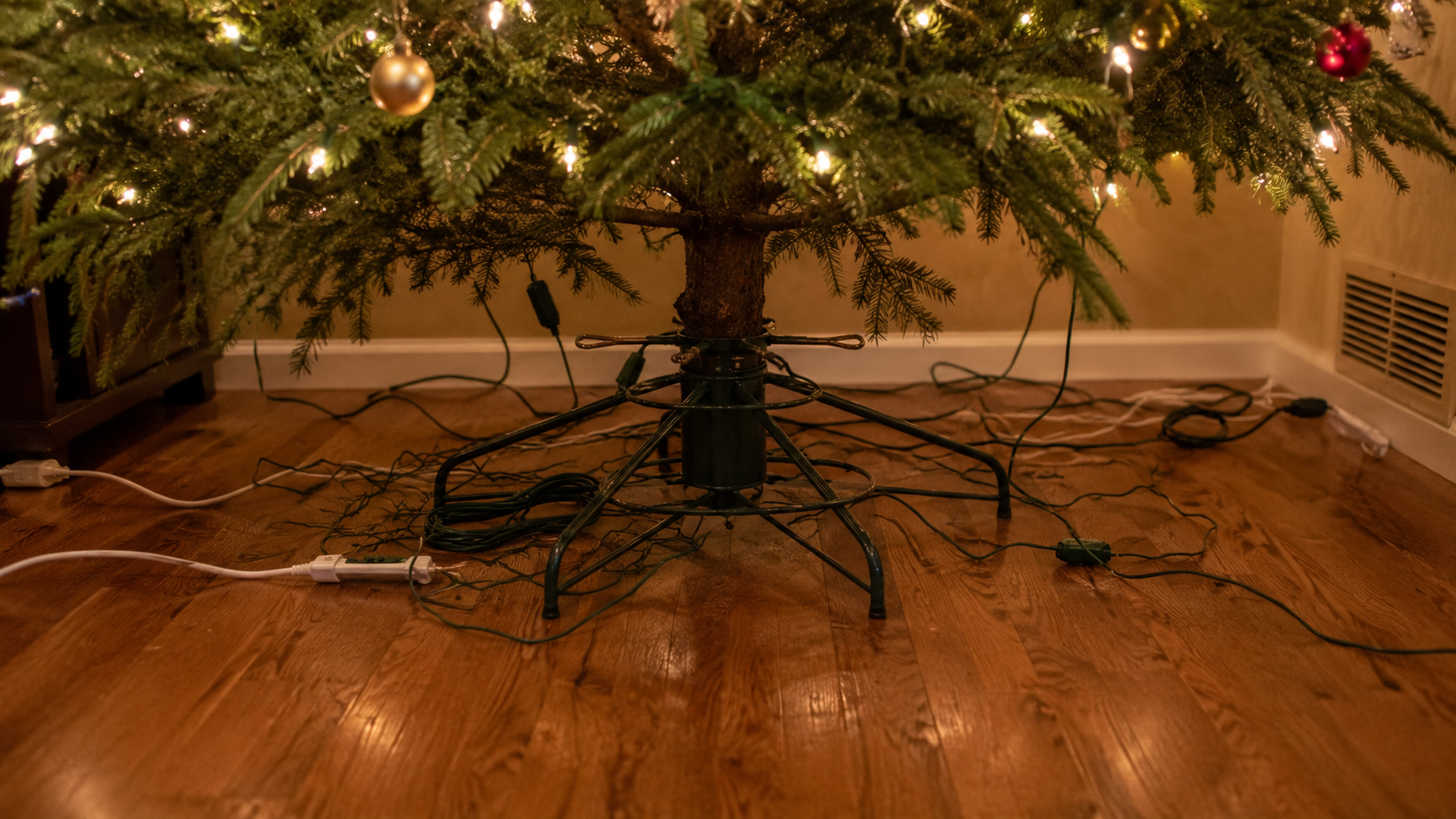 Suggested image: camera focused on the bottom of a Christmas tree, showing a messy metal tree stand, tangled electrical cords, and a floor that looks cluttered yet oddly empty before adding the tree skirt