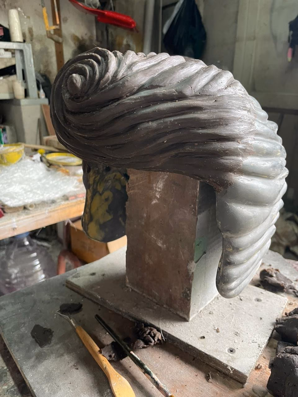 Hand-sculpted clay prototype of an Elvis pompadour helmet on a workshop stand showing material texture and detail.
