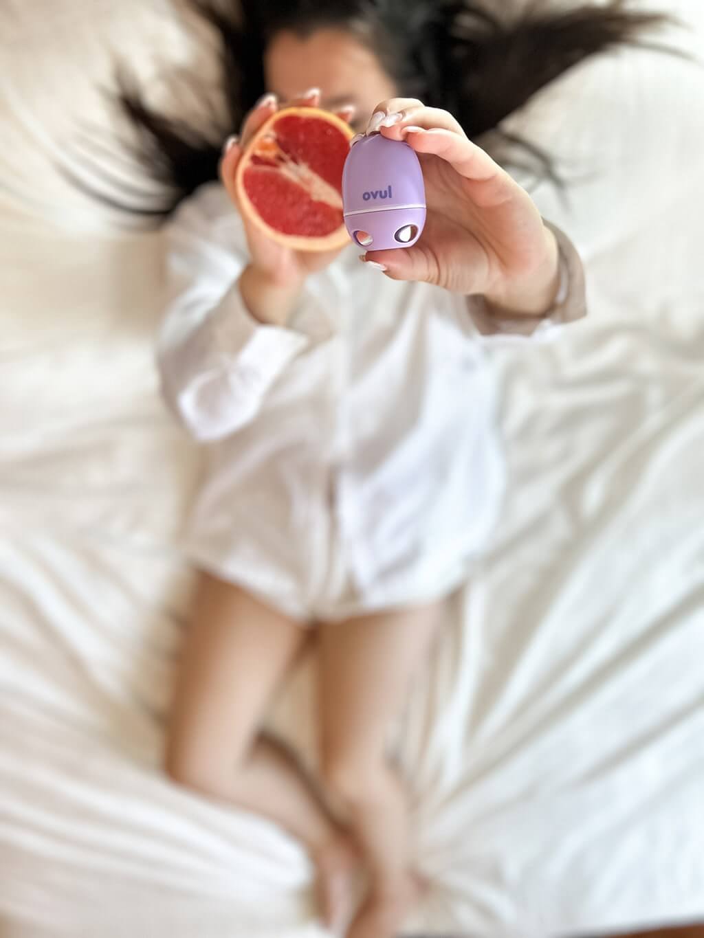 Woman holding Ovul AI app-based saliva fertility tracker and ovulation test next to a grapefruit half, laying on a white bed