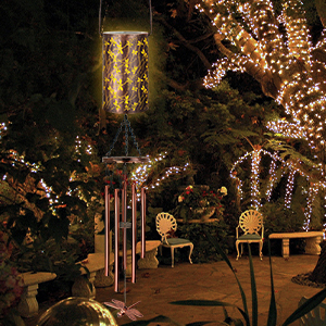Outdoor nighttime scene with decorative lighting. Metal wind chimes, illuminated lantern, and string lights in trees create a cozy atmosphere. Two chairs visible in background.
