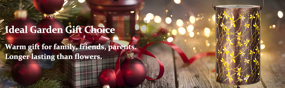 Cylindrical lantern with star cutouts emitting warm light, next to Christmas ornaments and wrapped gifts. Text suggests it's a garden gift alternative to flowers.