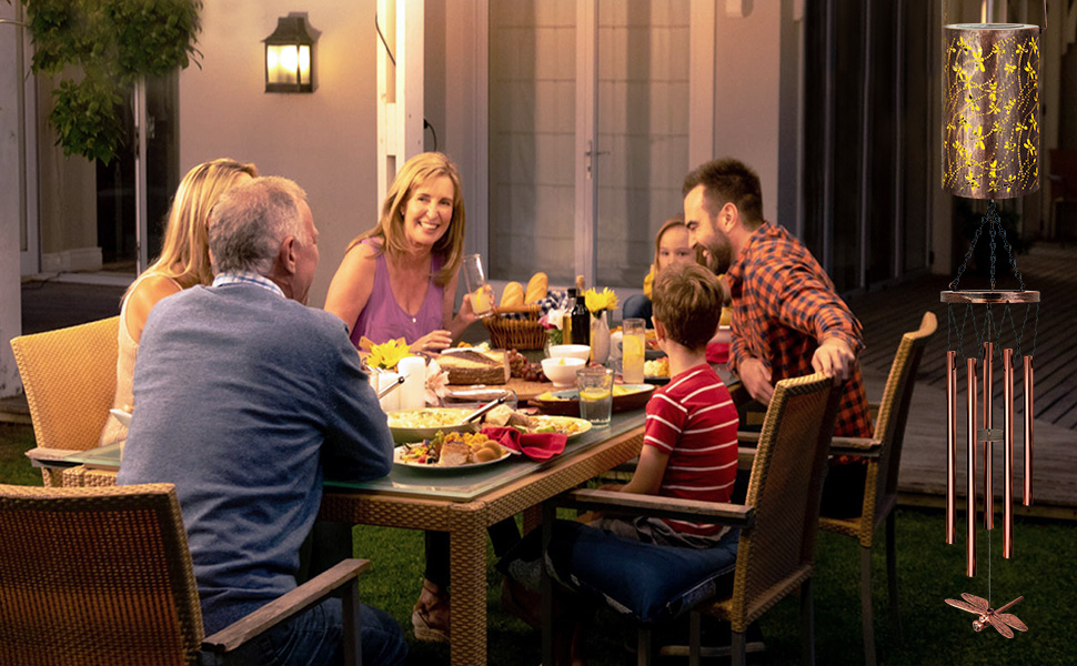 Outdoor patio dining scene with illuminated lantern and wind chime, showing people gathered around a table for a meal at dusk.