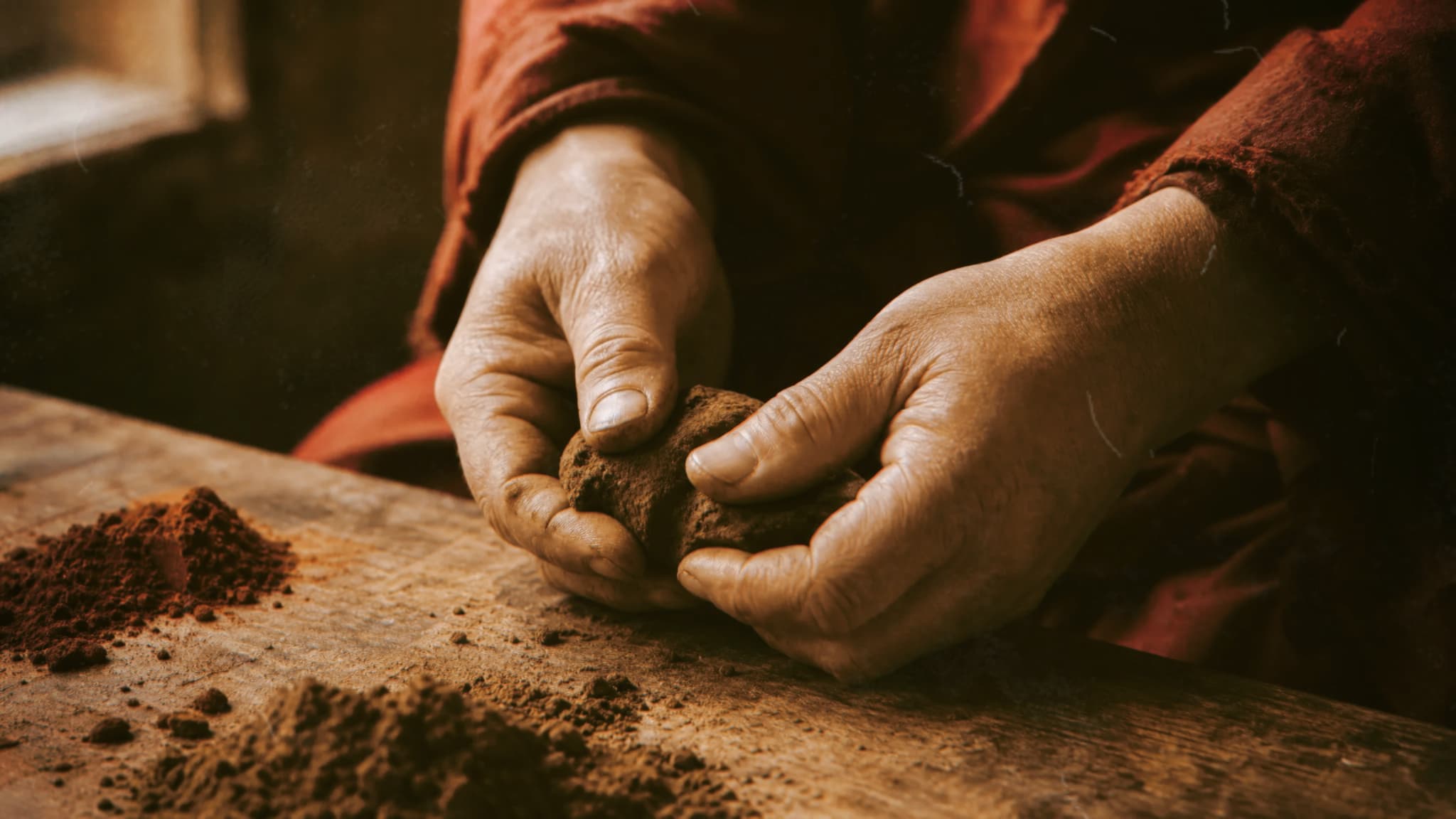 Close-up of hands shaping a brown mixture on a wooden work surface beside piles of powder.
