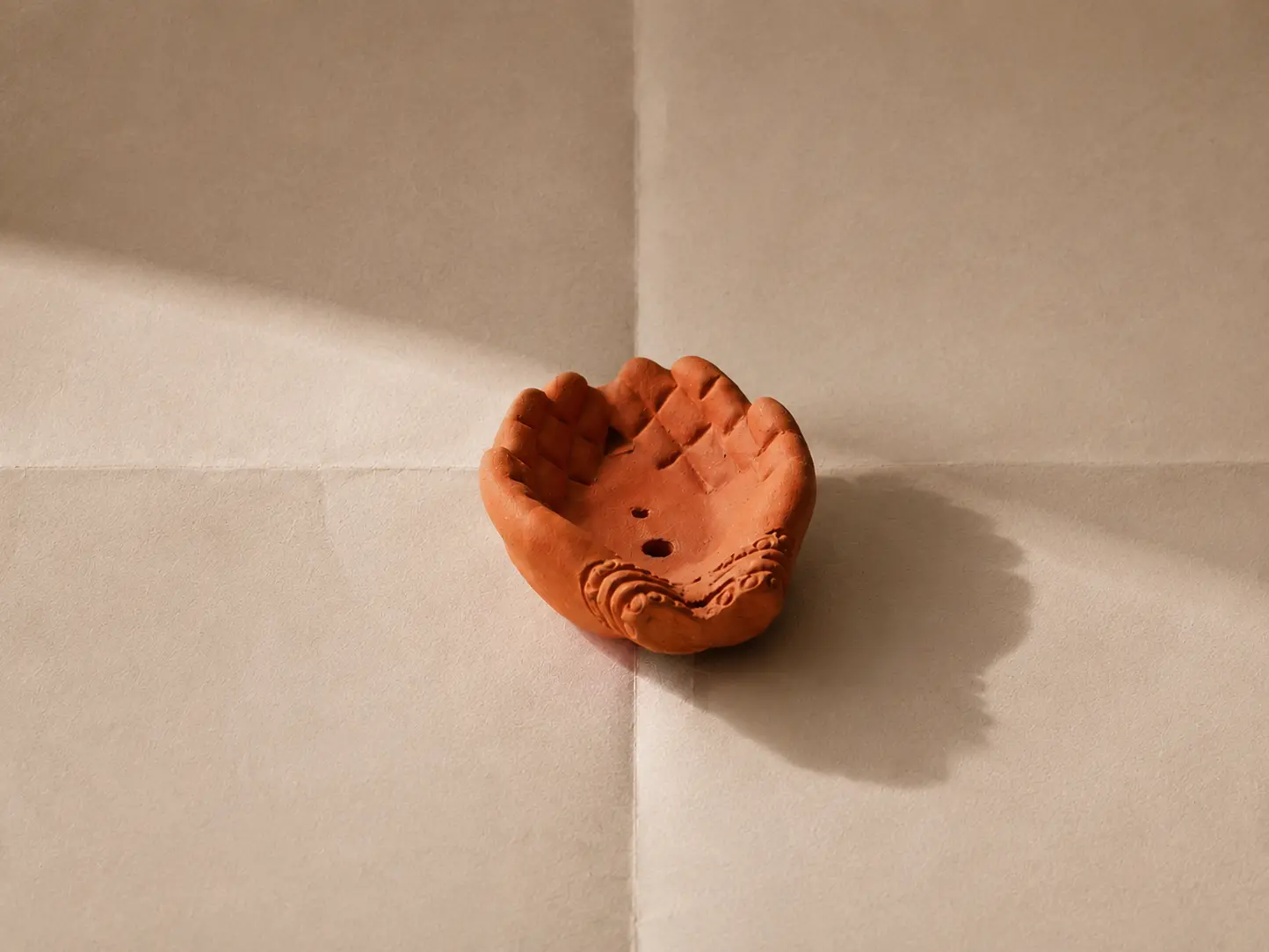 Pema hand-shaped red clay incense holder casting a shadow on a beige surface in sunlight