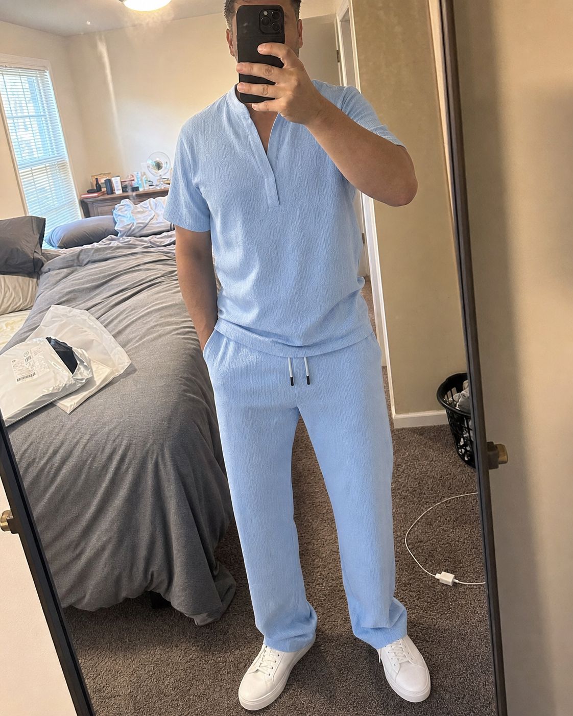 Phone mirror selfie of a midsize model wearing the Light Blue textured V-neck shirt and full-length pants set in a bedroom after unpacking