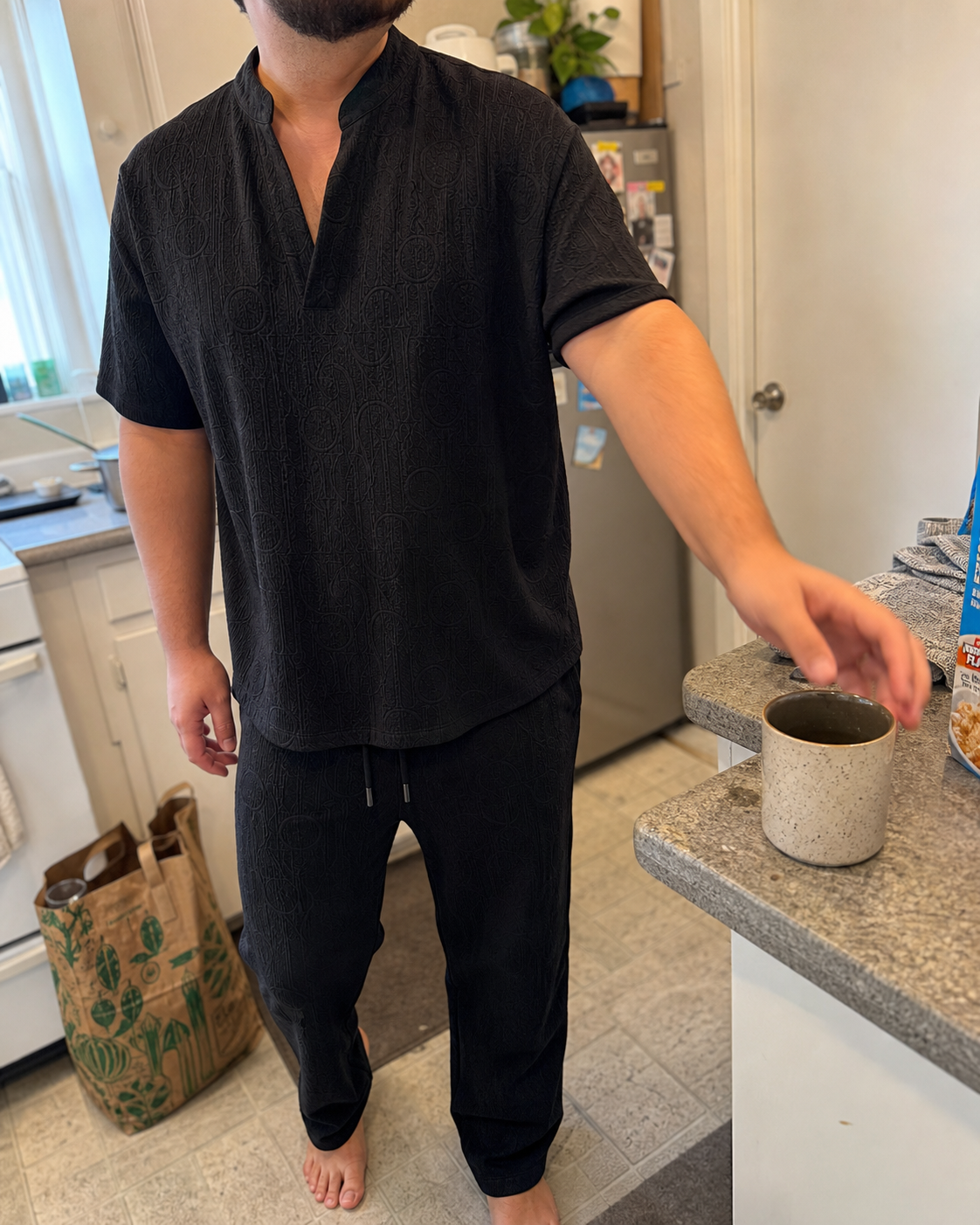 Candid three-quarter view of a stocky model wearing the Black textured V-neck shirt and full-length pants set while reaching for coffee in an apartment kitchen