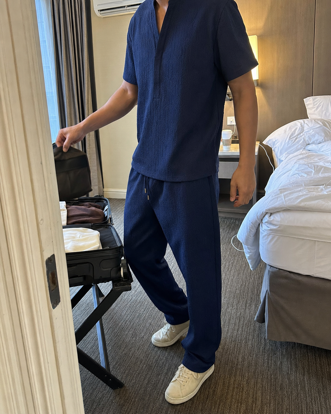 Three-quarter travel view of a tall model wearing the Navy Blue textured V-neck shirt and full-length pants set while unpacking beside an open suitcase