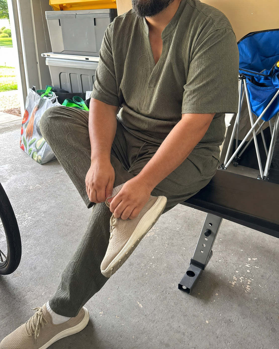 Seated three-quarter view of a plus-size model wearing the Army Green textured V-neck shirt and full-length pants set while tying a sneaker in a home garage