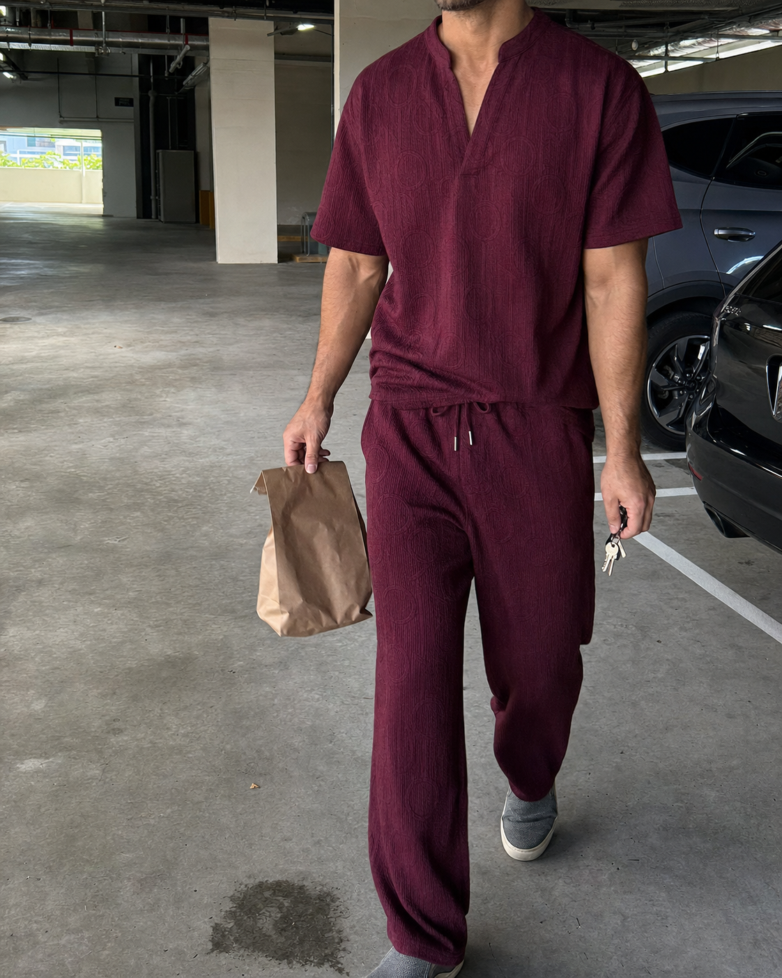 Candid walking view of an athletic model wearing the Wine Red textured V-neck shirt and full-length pants set while carrying a paper bag through apartment parking