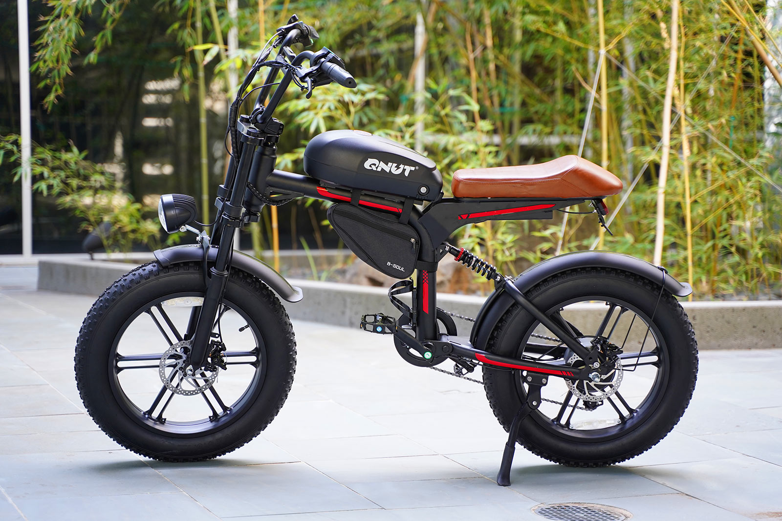 QNUT BK20 Electric Motorbike side view showing fat tires, full suspension, and retro-style brown saddle, photographed outdoors near bamboo.