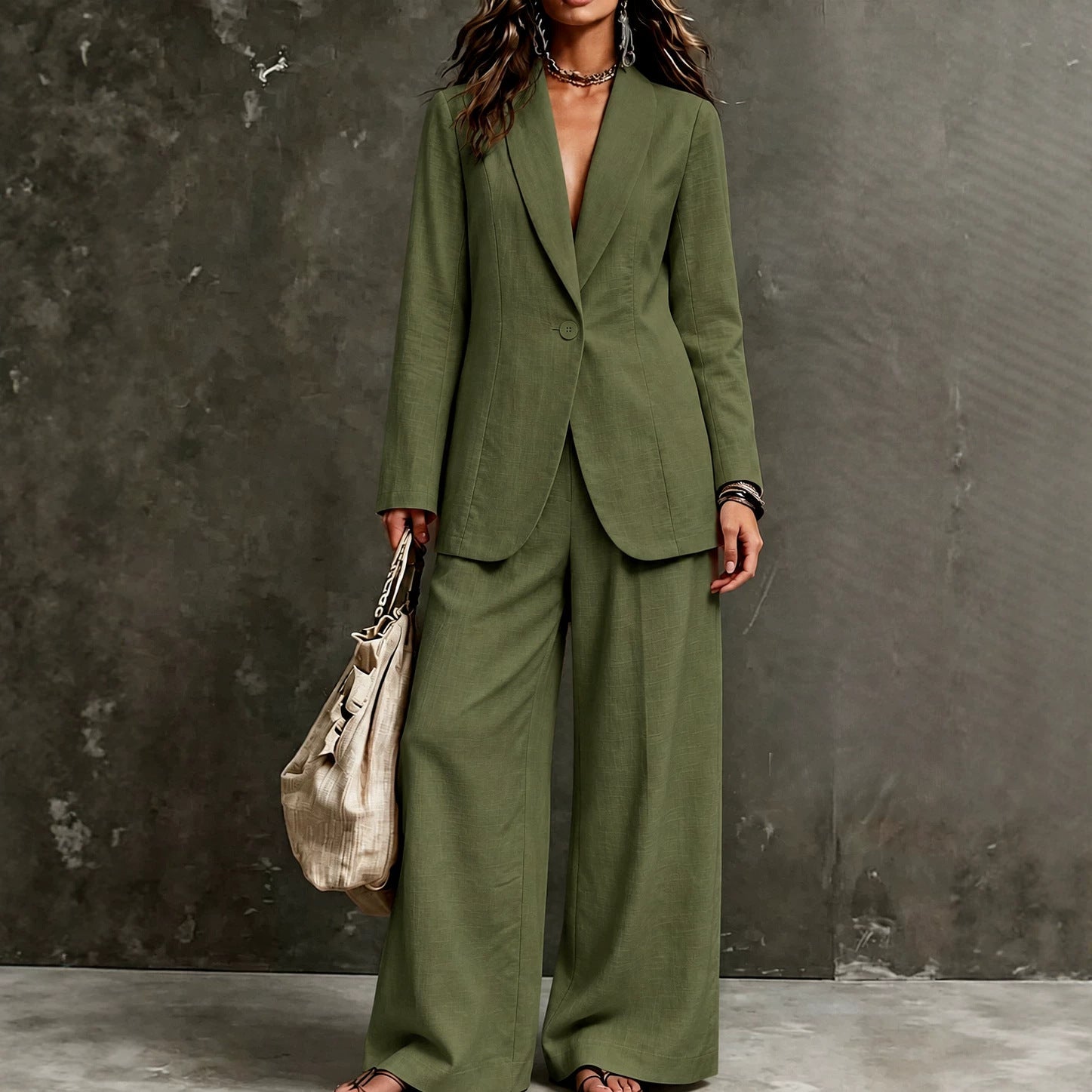 Women’s linen-look single-button notch-lapel blazer and wide-leg trouser suit set in Matcha Green – deep army olive green – full-length front standing view against warm grey concrete studio backdrop, fitted waist-suppressed blazer with notch lapel open V-neckline no inner layer, single tonal dark button at waist, front welt pockets, structured shoulders, full-length sleeves, hip-length hem, wide-leg straight full-length linen-look woven trousers, large beige woven tote bag in left hand, chunky layered chain necklace, dark beaded chandelier earrings, black bead bracelets stacked on right wrist, flat nude thong sandals